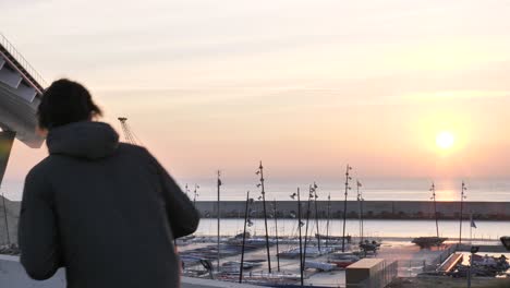 un joven con mascarilla en una scooter eléctrica se detiene frente al puerto de la ciudad para ver el amanecer, de mano