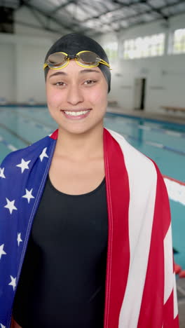 vertical video: smiling swimmer wearing swim cap and goggles, draped in american flag by pool