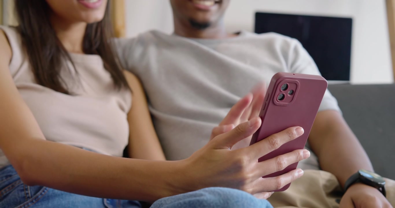 Coppia afroamericana su un divano grigio, la donna scorre un telefono rosa mostrando qualcosa, l'uomo appoggiato tocca l'orologio.