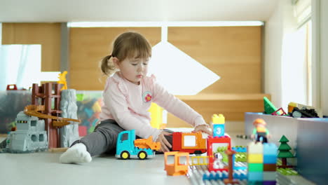 Cute-3-year-old-Caucasian-girl-playing-with-plastic-car-toys-on-a-floor-room-while-staying-at-home