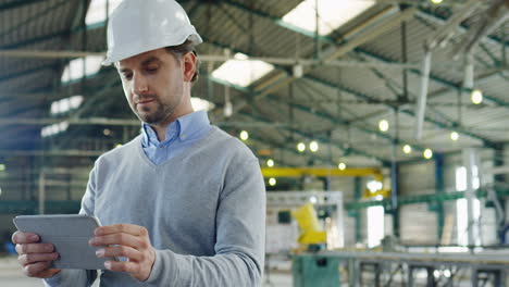 Trabajador-Masculino-Caucásico-Usando-Un-Casco-Usando-Una-Tableta-Y-Mirando-La-Cámara-En-Una-Fábrica