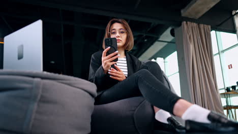 young brunette lady wearing glasses sits in bean bag chair. woman starts a phone call and looks at laptop. low angle view.