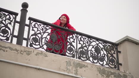 mujer con gorra roja y capucha descansa las manos en una intrincada barandilla de hierro bajo un cielo de invierno brillante, sonriendo pacíficamente mientras mira a la distancia, diseño elegante de la valla y atmósfera serena