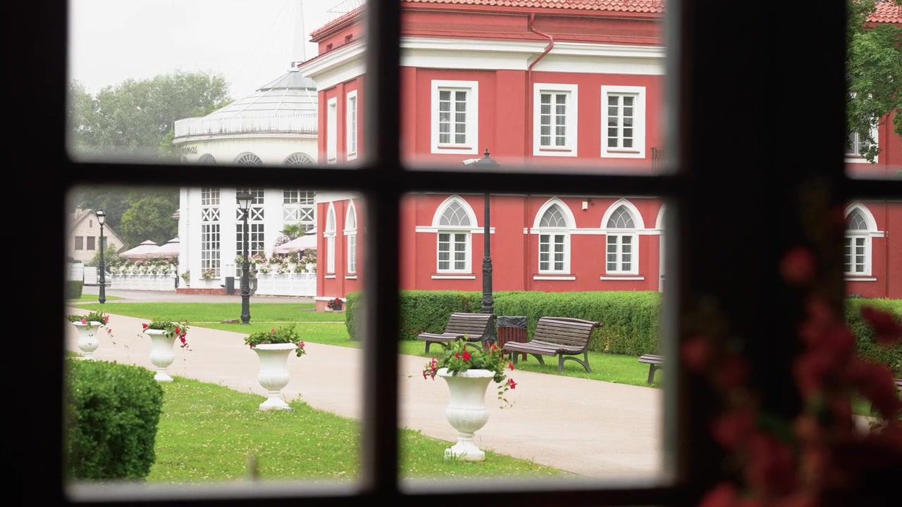 La morbida luce naturale del giorno illumina un tranquillo cortile al Castello di Raudondvaris, Lituania, incorniciato da una finestra con vivace architettura rossa, siepi verdi, panchine ed eleganti fioriere bianche