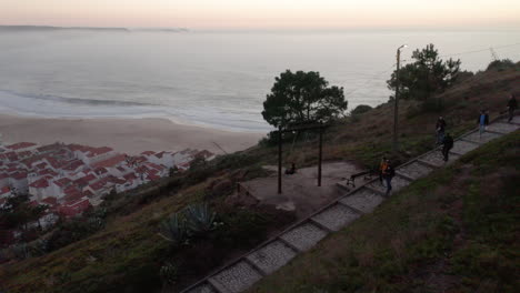 Turista-En-Sendero-Pavimentado-En-La-Cima-De-Una-Colina-Con-Vista-Al-Océano-Con-El-Casco-Antiguo-Durante-La-Puesta-De-Sol-En-La-Playa-De-Nazare,-Portugal