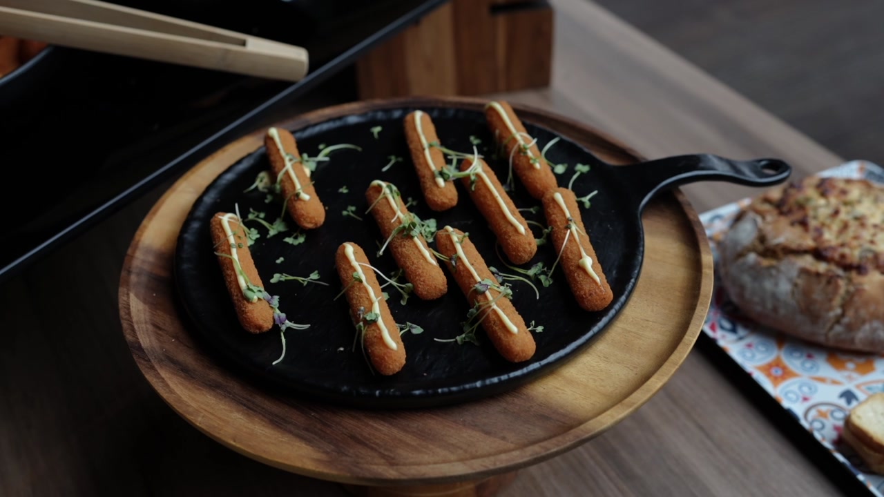 Fried croquettes drizzled with sauce and garnished with microgreens on a cast iron pan over wooden board, bread visible beside it