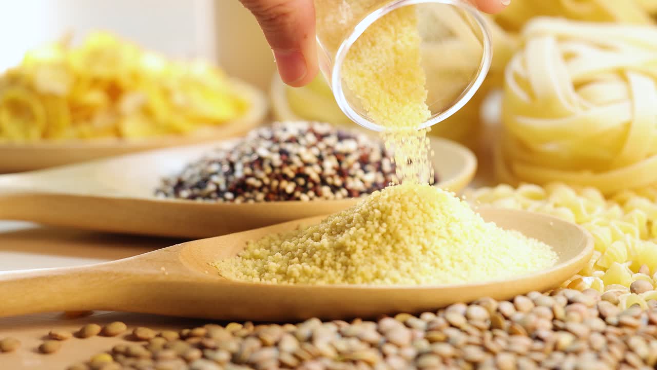A hand pours grains into a wooden spoon, surrounded by various grains and pasta, under warm lighting