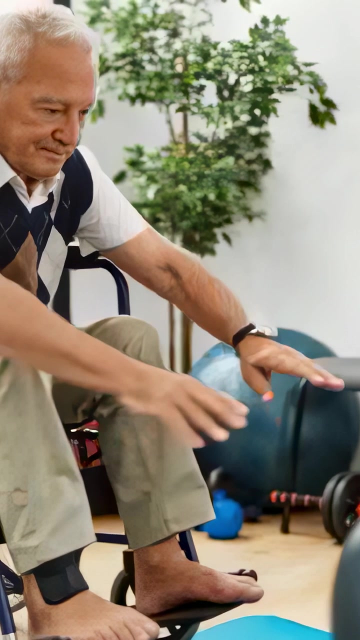 Vídeo vertical: Idoso em cadeira de rodas se movendo e alcançando uma bola azul em uma sala de reabilitação, com espaço para texto.