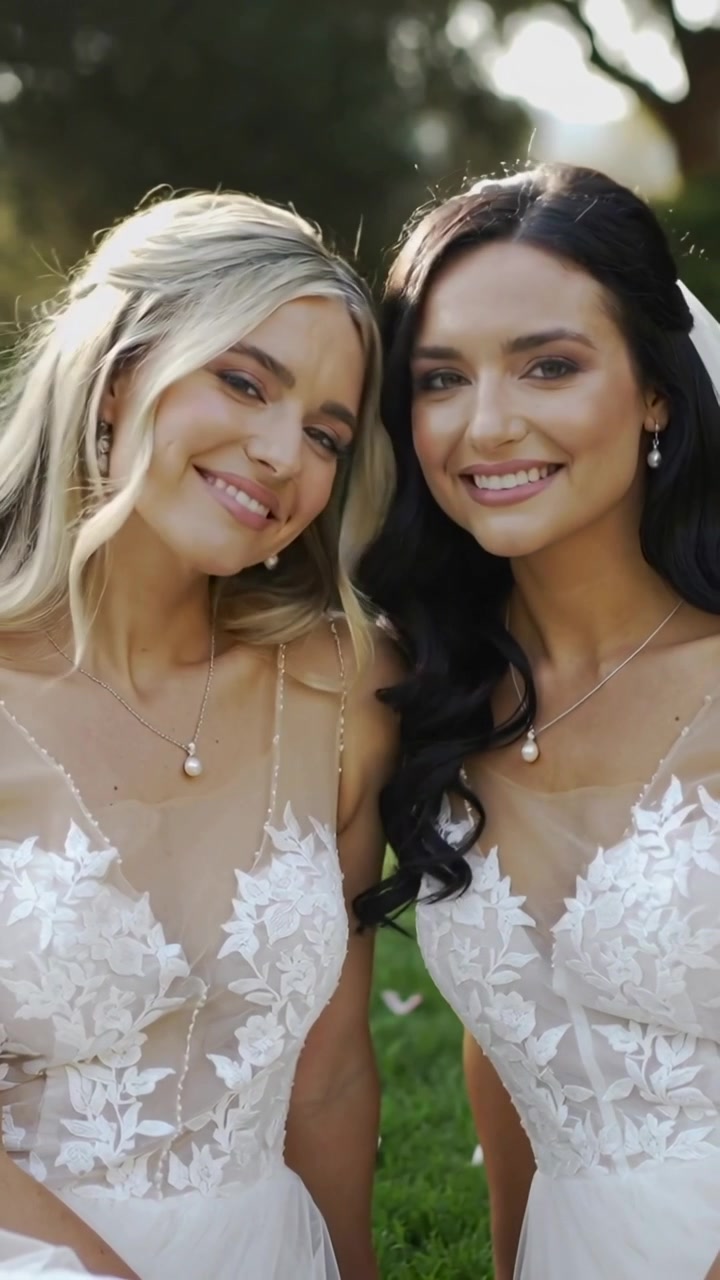 Vidéo verticale : Deux mariées en robes de dentelle sourient et penchent la tête en posant dans un jardin.