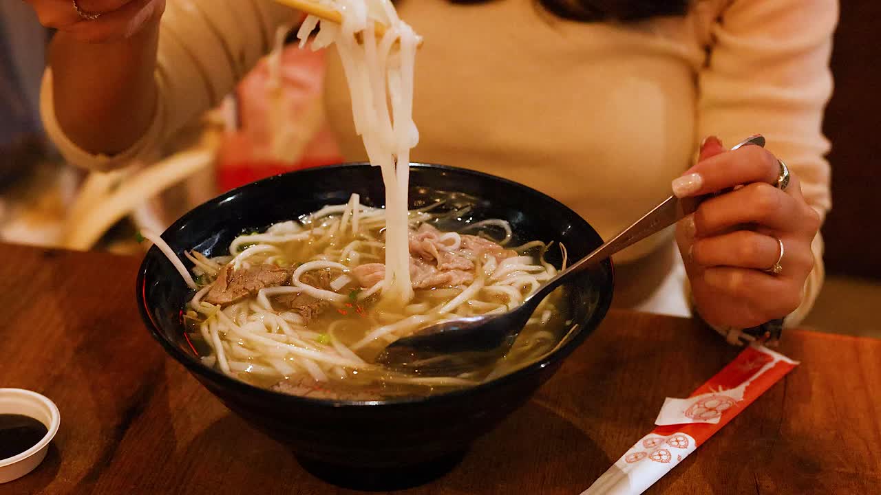 A person savoring a bowl of Vietnamese pho with chopsticks and a spoon in a cozy setting