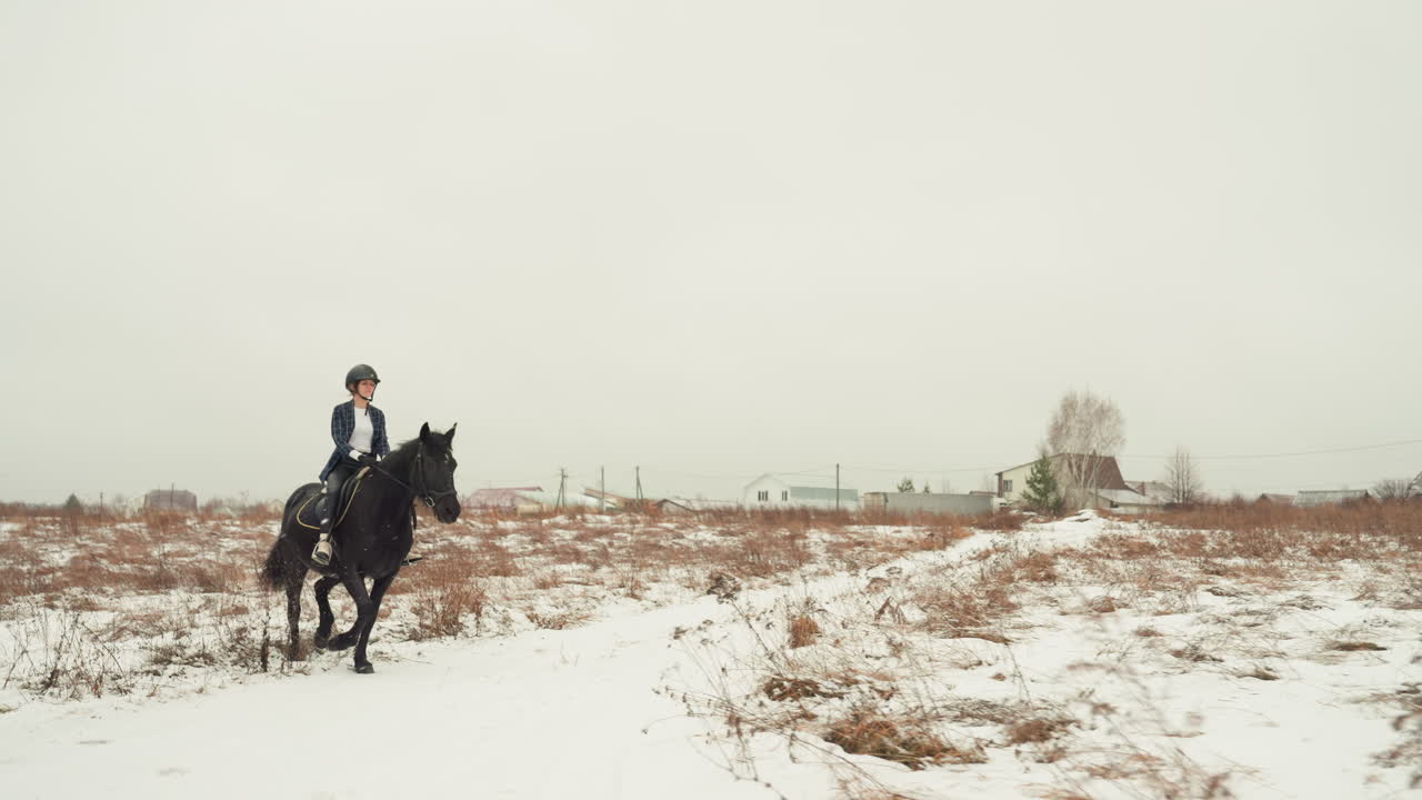 Una donna cavalca un cavallo nero attraverso un paesaggio innevato. Sotto un cielo grigio, la cavallerizza percorre tranquillamente un sentiero rurale innevato. La donna cavalca con attenzione il suo cavallo scuro attraverso la campagna invernale con case sparse.