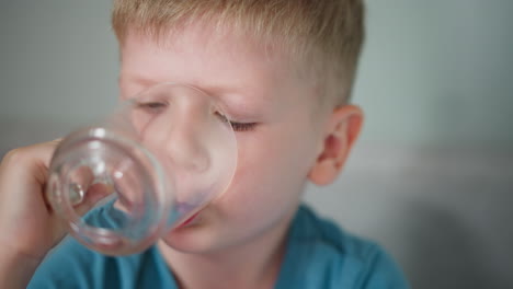 close up of little boy with blond hair drinking water from clear glass, sitting on bed in cozy home setting, wearing blue shirt, focused on hydrating, refreshing moment of childhood
