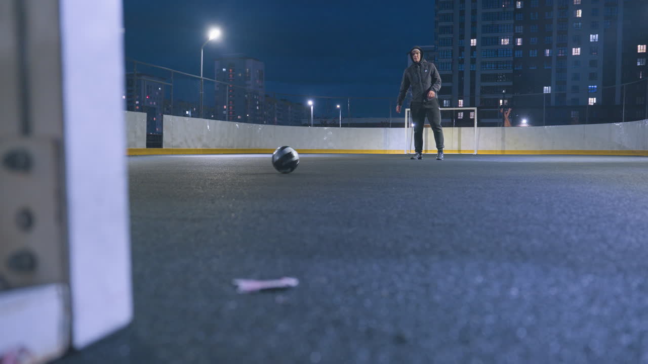 Athlete kicking soccer ball toward goal post during intense night training session on urban outdoor field, ball rolls out of post while city lights and distant skyline create a vibrant backdrop