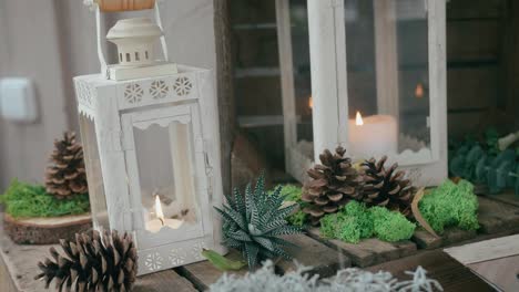 Decorations-made-with-candles-inside-white-lanterns,-small-plants-and-pinecones,-on-a-wooden-surface