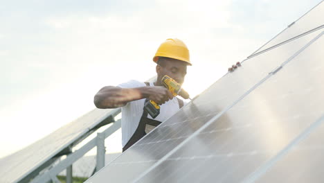 Afroamerikanischer-Mann-In-Spezieller-Uniform-Und-Schutzhelm,-Der-Ein-Solarpanel-Repariert