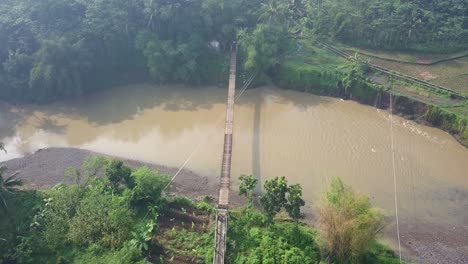 Vista-Aérea-De-Drones-Del-Puente-Colgante-Sobre-El-Río-Con-Cruce-De-Motocicletas-En-La-Mañana-Nublada-Con-Rayos-Solares,-Java-Central,-Indonesia