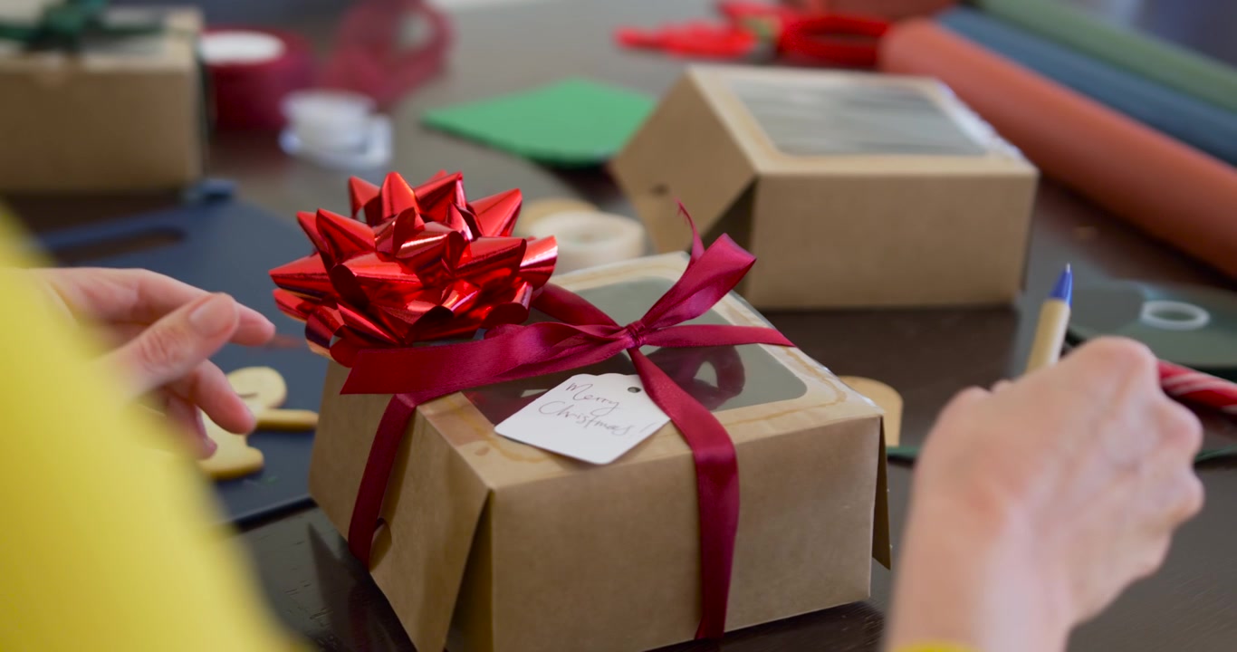 Donna che finisce un regalo di Natale, scrivendo con una penna blu e lisciando il nastro sotto un fiocco rosso al tavolo