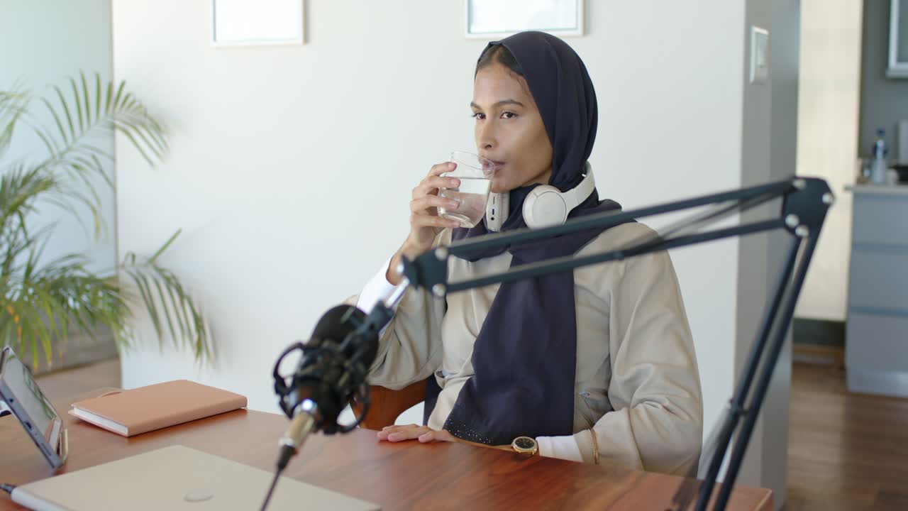 Kvinde pauser podcast ved skrivebordet, rækker ud efter et klart vandglas for at hydrere, drikker, kigger kort på mikrofonen.