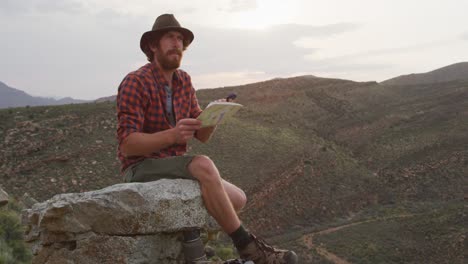 Caucasian-male-survivalist-sitting-on-mountain-peak-in-wilderness,-holding-map-and-compass