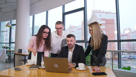 bearded man in suit sits in front of laptop and talks. his colleagues gathered around him and listen to the speaker.