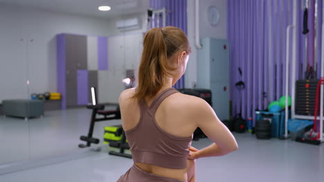 vista trasera de una mujer en una alfombra de yoga púrpura realizando un estiramiento profundo, con un gran espejo que refleja su postura y el equipo de gimnasia circundante