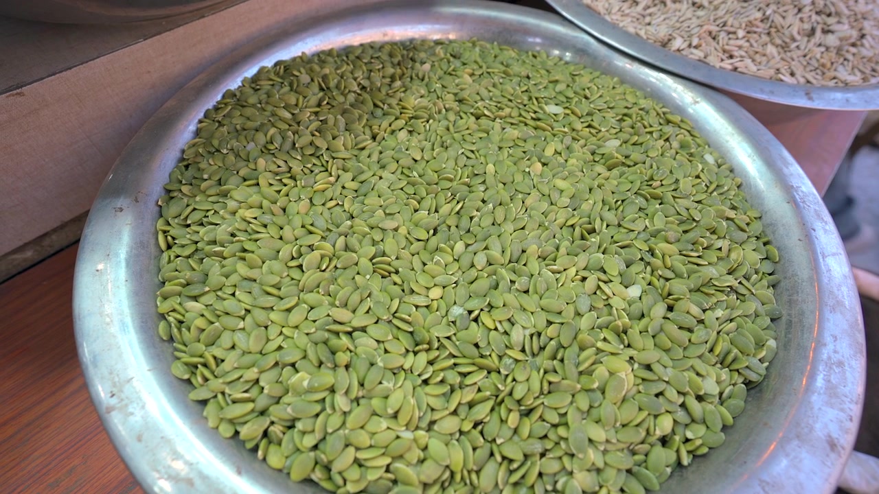Una ripresa statica ravvicinata dall'alto di una grande ciotola rotonda di alluminio piena di vivaci semi di zucca crudi o pepite in un'esposizione di mercato in India.