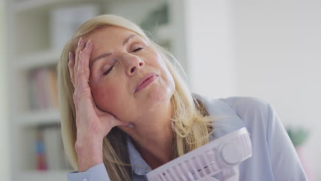 mujer madura en la menopausia que tiene sofocos en casa refrescándose con un ventilador eléctrico