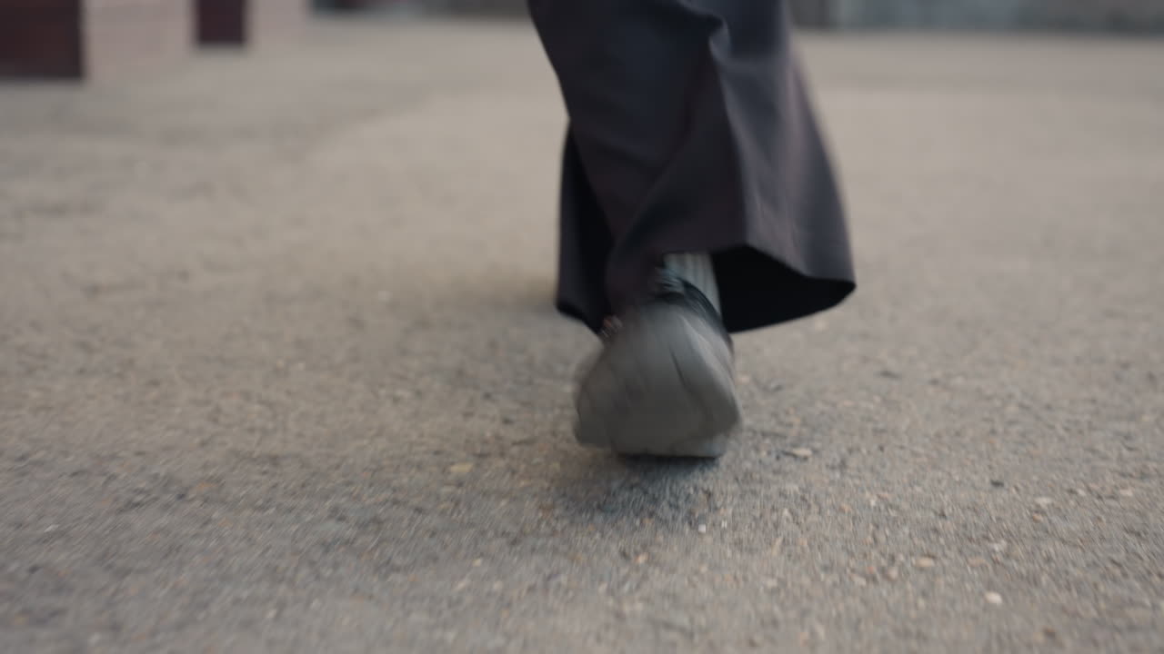 Primer plano de una persona manteniendo las piernas alineadas mientras camina hacia la cámara sobre un suelo arenoso; un zapato negro marca el paso firme, el dobladillo del pantalón roza el asfalto y el movimiento sugiere un desplazamiento urbano resuelto.