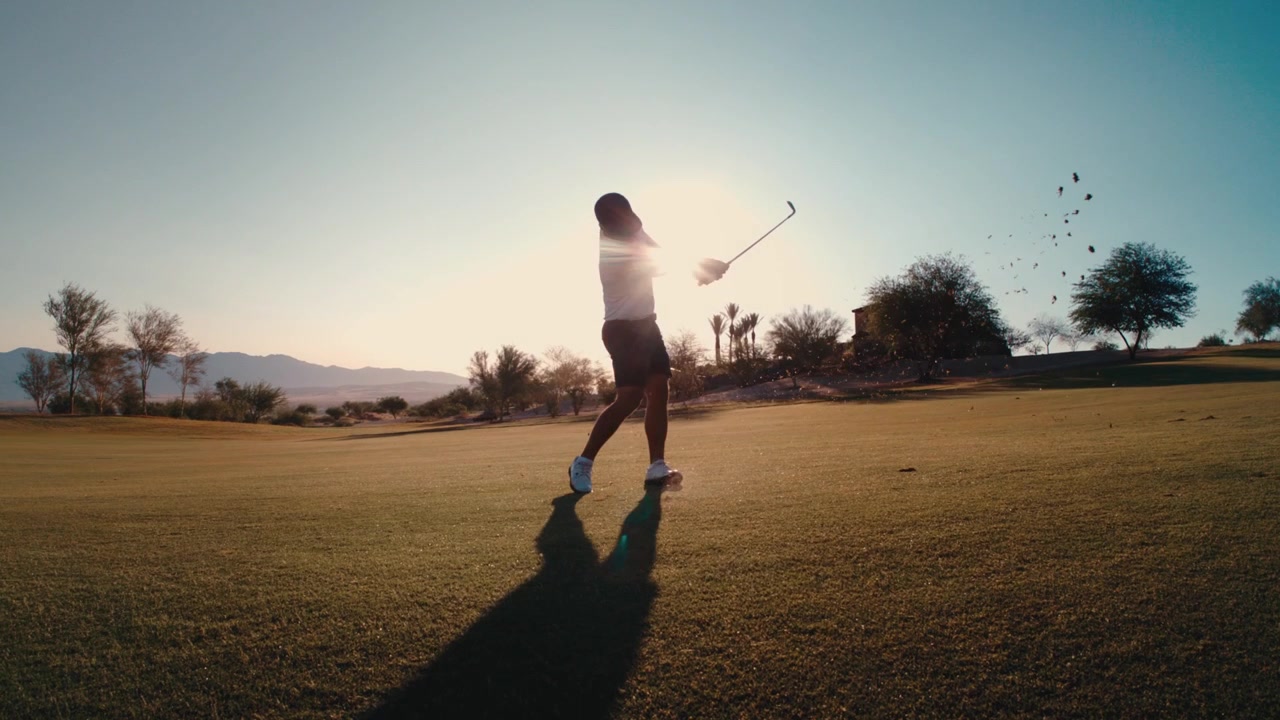 Golfer hits wedge pitch at golden hour in the desert, silhouetted against a sun flare through filmed in slow motion,