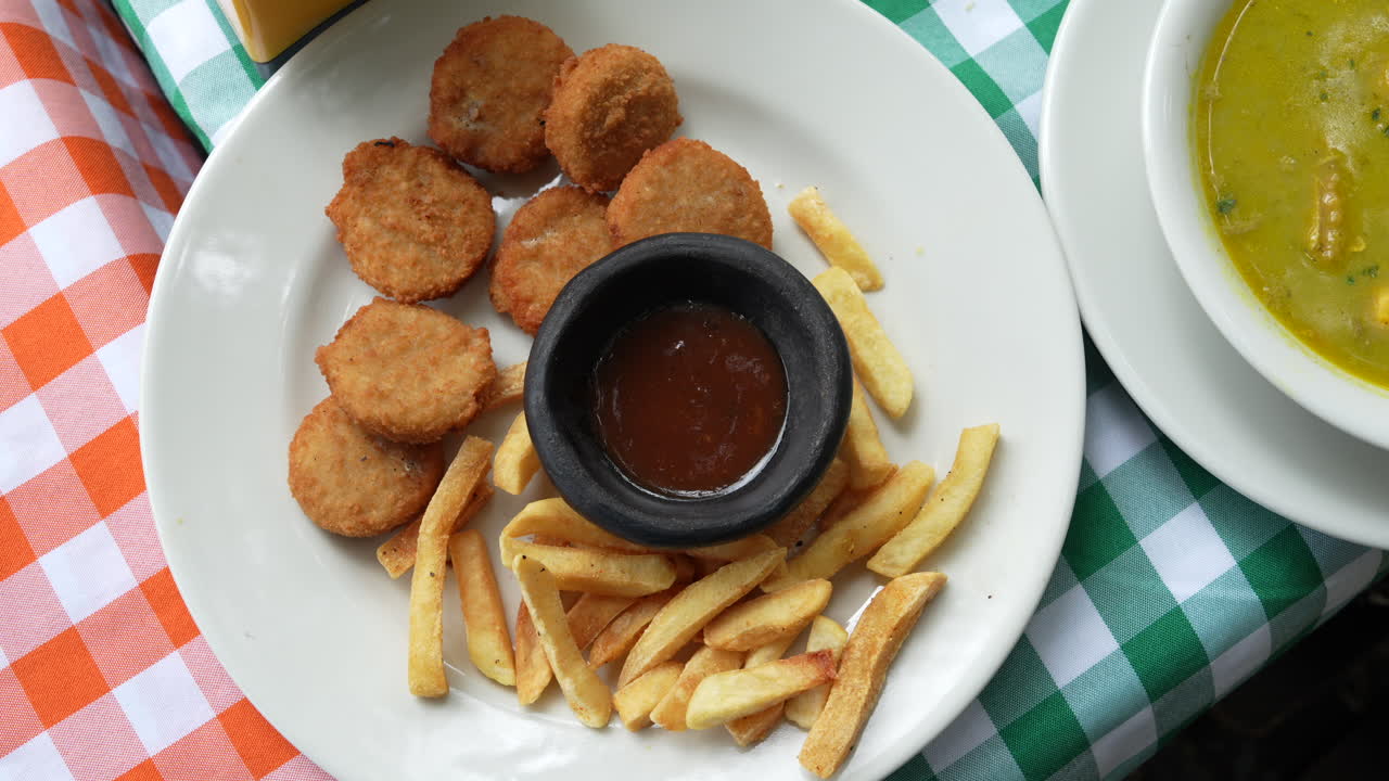 Deliciosa foto em close-up de nuggets crocantes servidos com batatas fritas douradas e um rico molho para mergulhar em uma vibrante toalha de mesa xadrez. Perfeito para amantes da gastronomia e entusiastas culinários