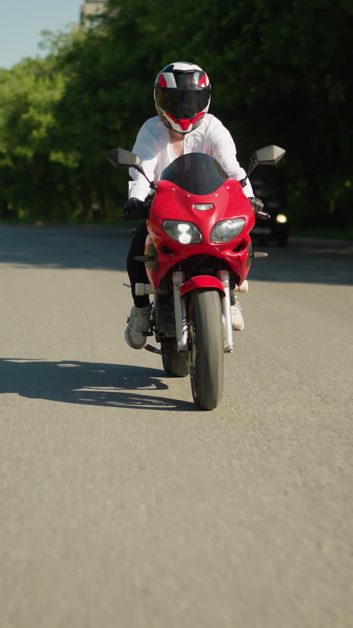 una motociclista femenina con casco viaja rápido en una bicicleta eléctrica roja a lo largo de una carretera, el fondo muestra un borrón de árboles verdes exuberantes y un coche que se acerca por detrás