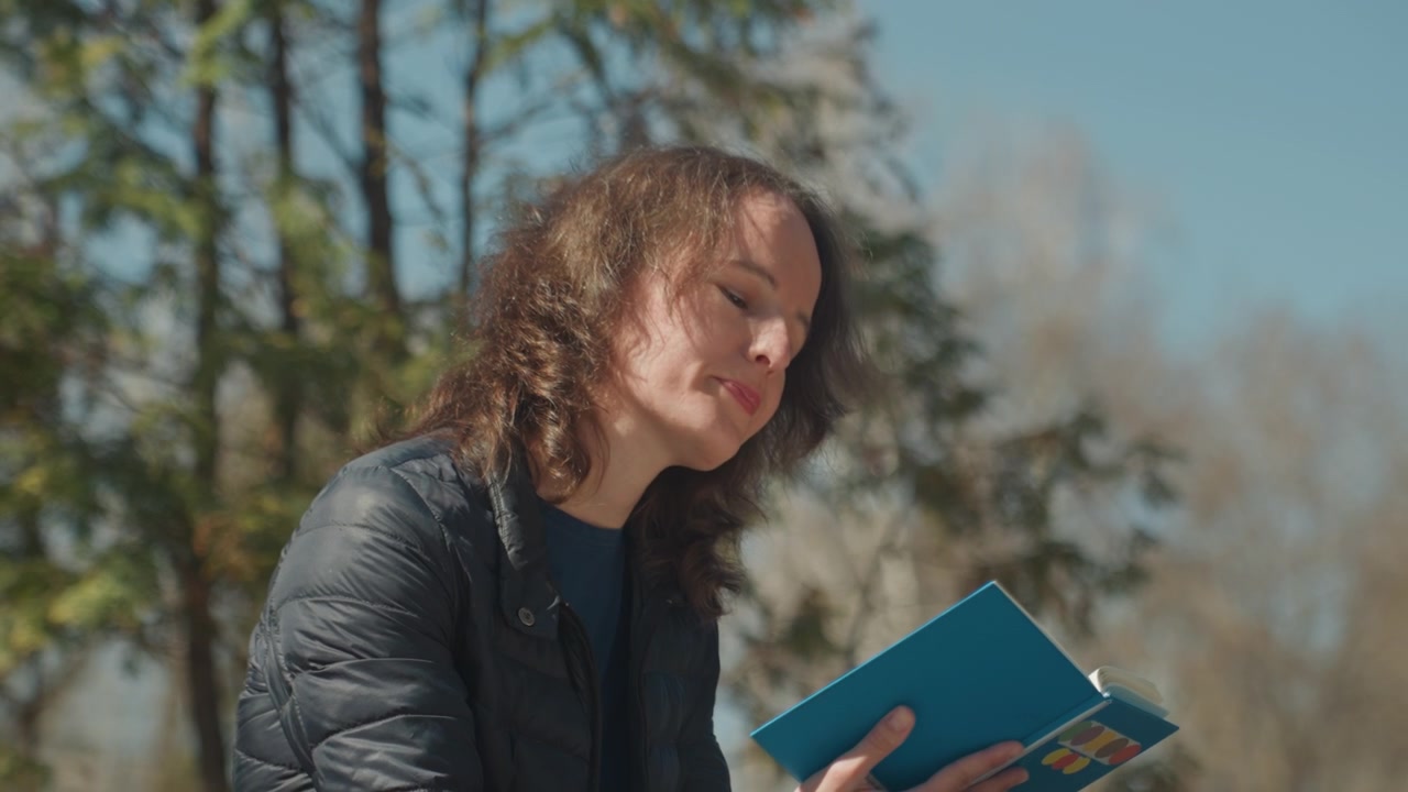 Primo piano di donna che legge un libro blu all'aperto con uno sfondo di alberi e cielo. Il libro è aperto, tenuto nella mano sinistra, la giacca è parzialmente chiusa con la cerniera. Una luce naturale soffusa illumina la scena.