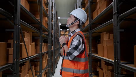 vista lateral de ingeniero masculino asiático con casco de seguridad reza por algo mientras está de pie en el almacén con estantes llenos de bienes de entrega
