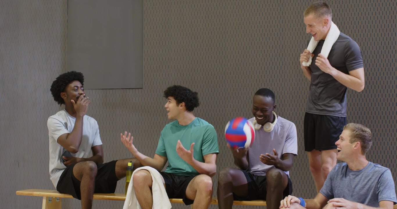 Diverse männliche Teamkollegen kommen an, umarmen sich, spielen sich einen Volleyball zu und unterhalten sich nach dem Training auf der Turnhallenbank.