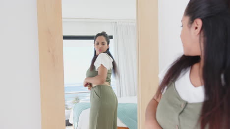 Looking in mirror, asian woman in green jumpsuit adjusting her outfit, contemplating style, at home