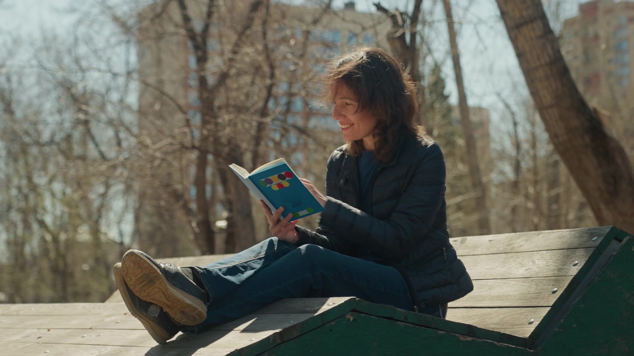 Close up of woman reading book outdoors on wooden structure with trees and buildings in background. Book held open casual posture sunlight casting shadows on wood. Scene calm with natural