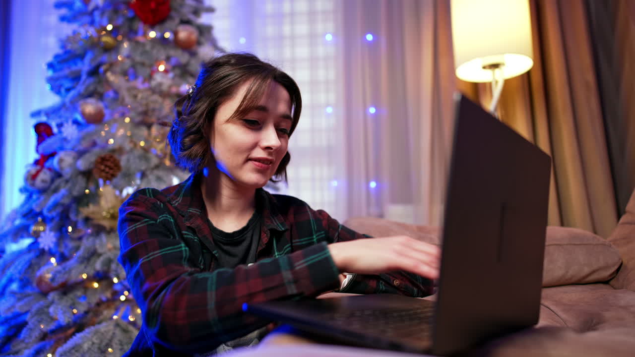 Cozy night by the tree. A young woman works on her laptop by a decorated Christmas tree in a warm living room during the holiday season