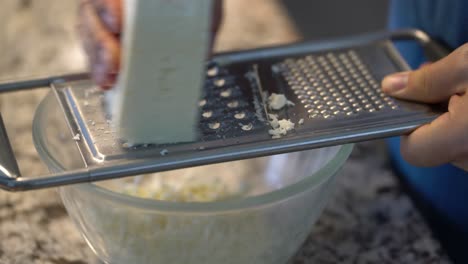 close up of hand grating block of feta on cheese shredder in home kitchen