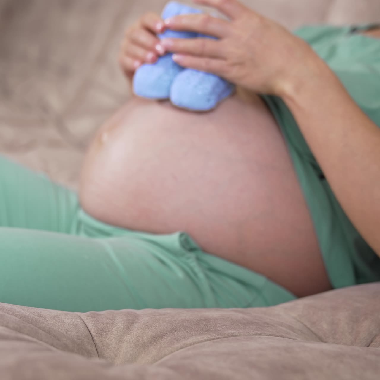Pregnant woman with bare belly at home. Beautiful knitted blue baby socks on a sofa. Future mother stroking her belly. Pregnancy concept