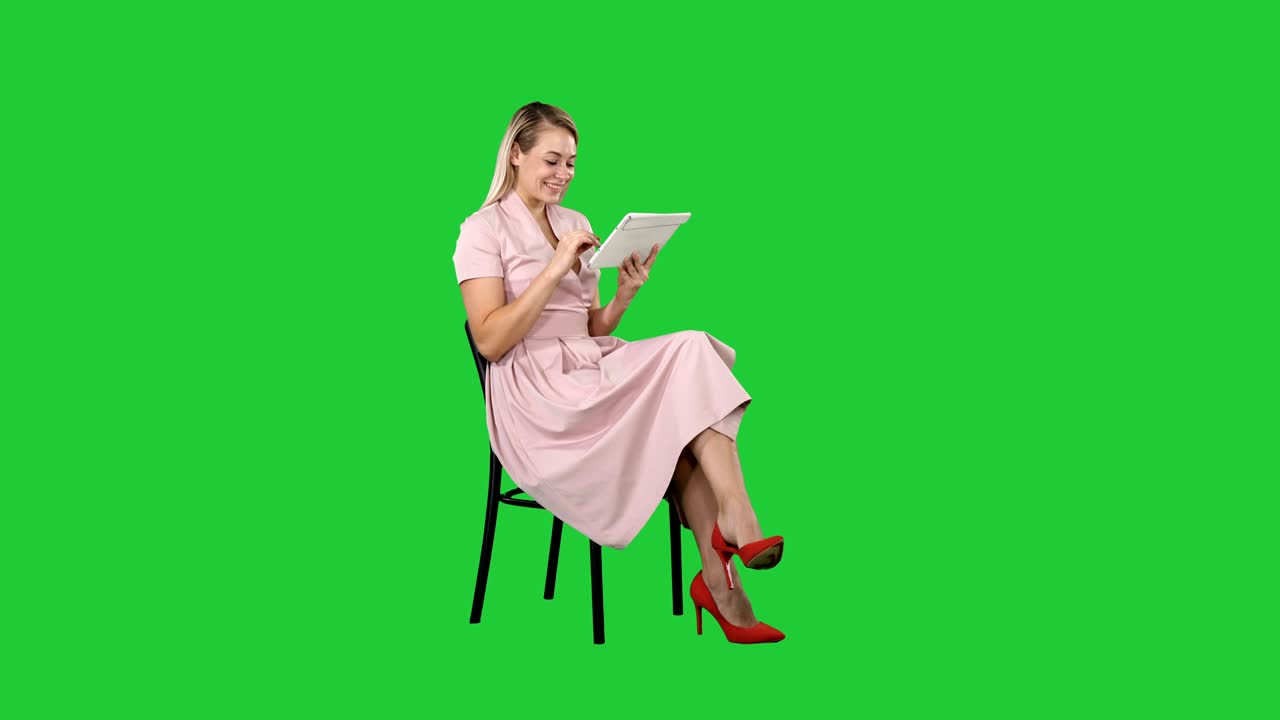 핑크색 드레스를 입은 재미있는 예쁜 젊은 여성이 의자에 앉아 녹색 화면에서 태블릿을 사용하여, 크로마 키