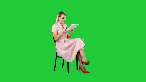 funny pretty young woman in pink dress sitting on a chair and using tablet on a green screen, chroma key