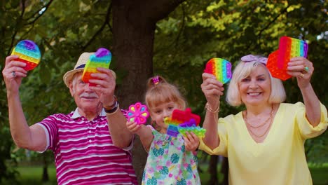 Senior-grandmother-grandfather-with-granddaughter-holding-anti-stress-pop-it-toy-games-in-park