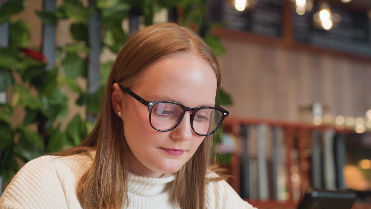 Close up of adult woman wearing glasses and white sweater looking at phone with soft bokeh background inside cozy cafe filled with warm lights and indoor plants