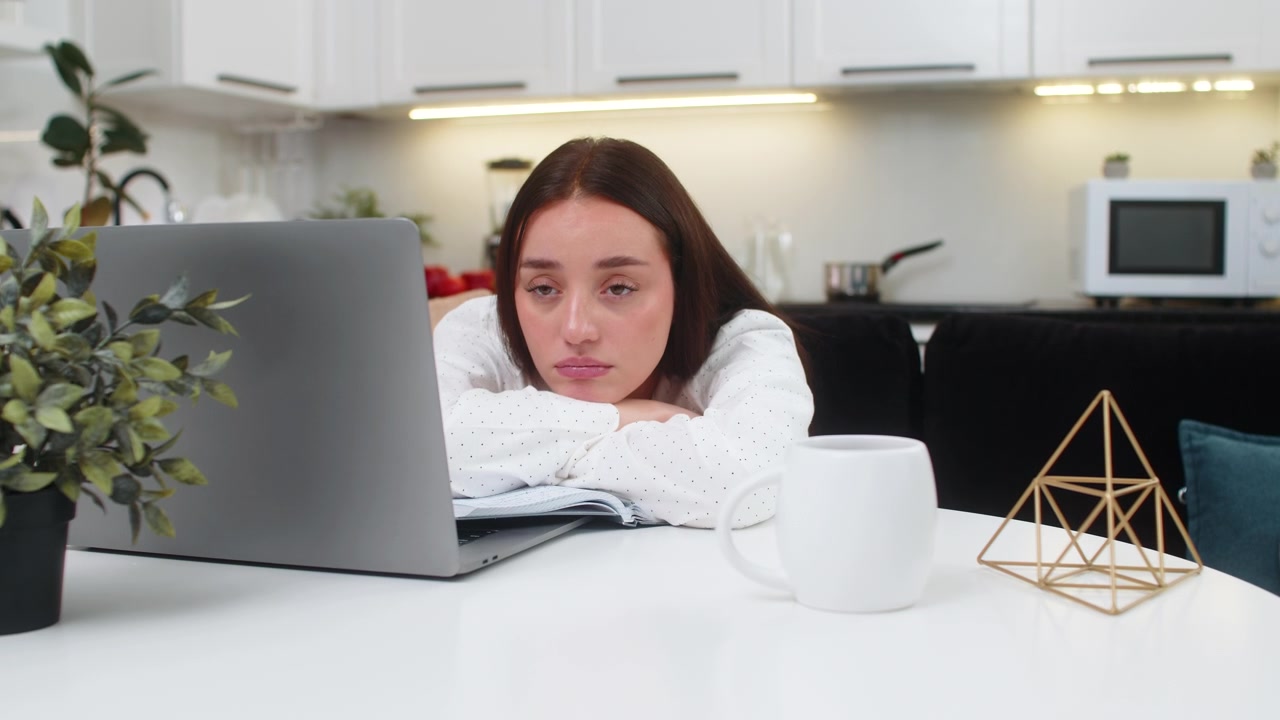 Giovane donna prende appunti sul quaderno con laptop, annoiata, bassa energia, demotivata, burnout, routine, stanchezza.