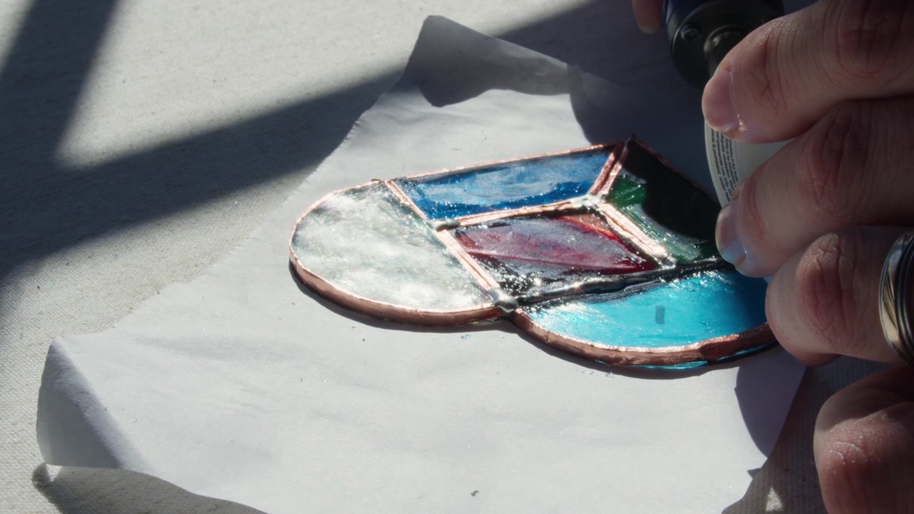Ein Student lernt, wie man ein Buntglas-Kunstwerk herstellt, indem er Draht auf ein Design aus geschnittenen Glasformen schmilzt. Das Glas wird dann fixiert und der Lehrer weist auf ein Detail hin.