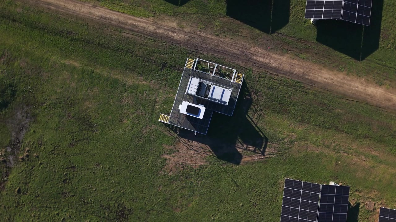 Droneopptak viser en solbelyst solcellepark i The Basin, Melbourne, som fremhever elegante paneler og en liten bygning omgitt av frodig gress og grusveier, og understreker fornybar energi.
