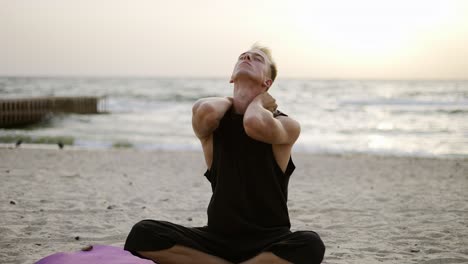 un joven estira su cuello mientras hace yoga durante el amanecer en la playa. ejercicios deportivos al aire libre