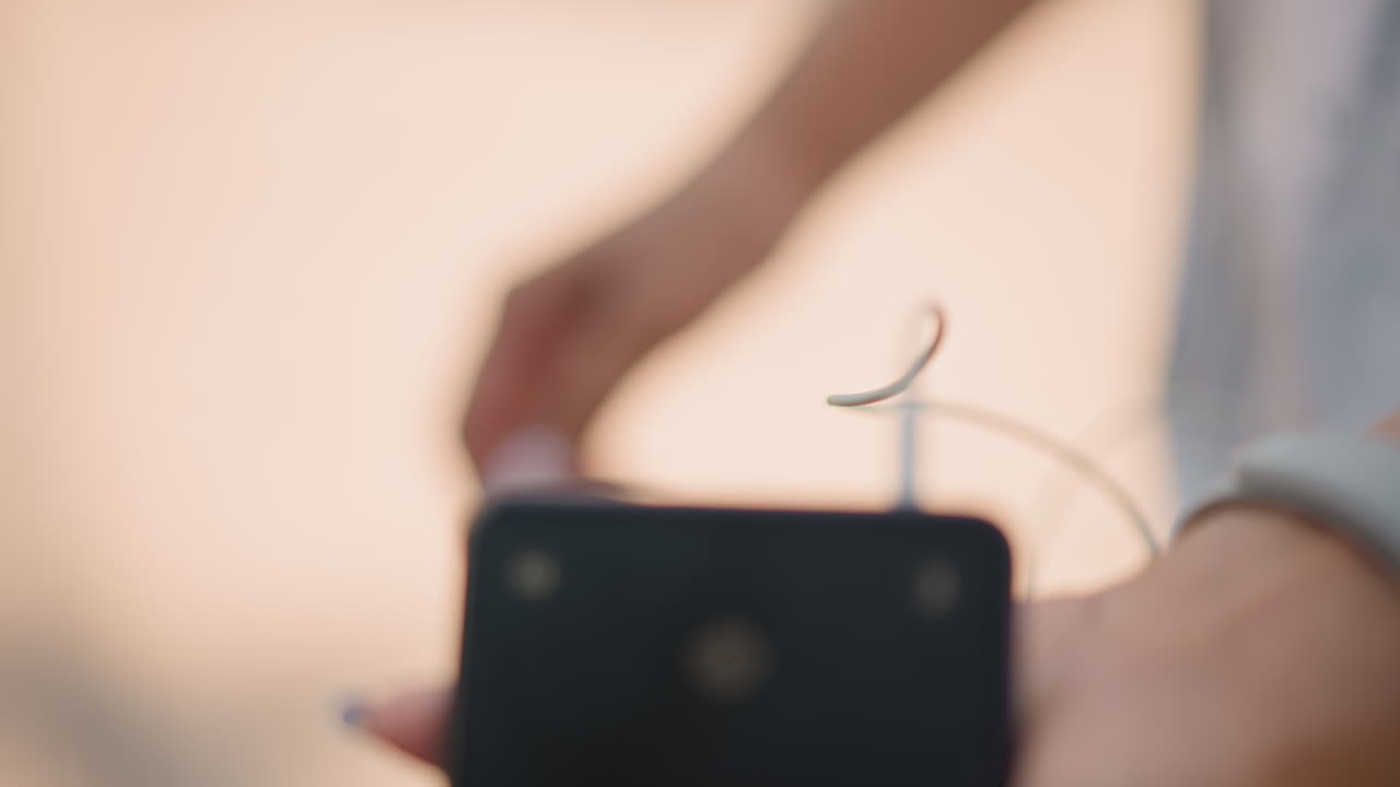 primo piano di mani che inseriscono un auricolare nello smartphone, runner che indossa uno smartwatch bianco, bokeh caldo al tramonto, movimento attento delle dita che inseriscono il jack .mm nella porta, cavo avvolto intorno al telefono, fitness soft