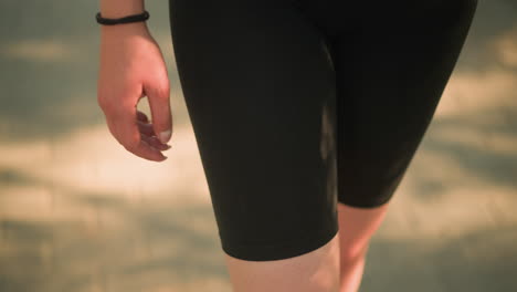 partial leg view of lady in black short leggings, wearing black hand bangle, walking outdoors under warm sunlight, with blurred background featuring soft shadows on ground