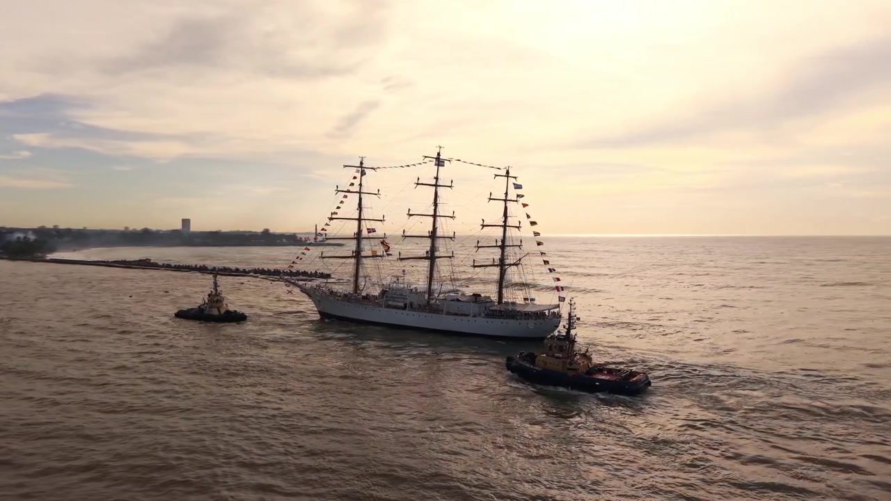 Ripresa aerea della nave scuola Fragata Libertad scortata da rimorchiatori verso il porto di Santo Domingo durante l'ora d'oro, evidenziando la logistica marittima e il patrimonio nautico